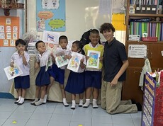 Mehrere Kinder stehen in einem Klassenzimmer und halten bemalte Blätter hoch; ein junger Erwachsener kniet neben ihnen.