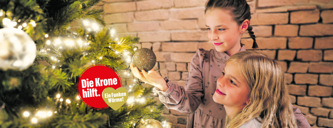 Zwei Mädchen beim Christbaum - und ein Logo "Die Krone hilft. Ein Funken Wärme"