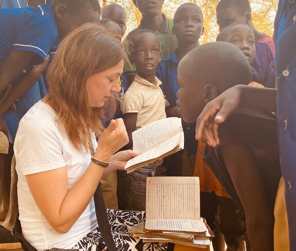 Natürlich — hier ist der überarbeitete Alternativtext als ausgewogener Mix beider Varianten:  > **Alternativtext:** > Die Bereichsleiterin Alexandra Blattnig-Rull der Auslandshilfe der Caritas Kärnten sitzt bei einem Projektbesuch und liest in einem Heft, während sie von Kindern der Schule umringt ist und mit ihnen spricht.