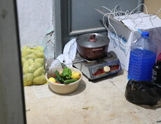 Kochtopf auf einem kleinen Gaskocher neben Gemüse, Wasserkanister und Haushaltsgegenständen.