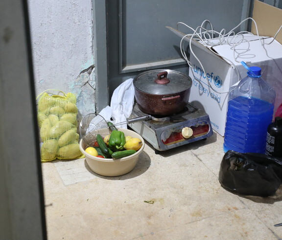 Kochtopf auf einem kleinen Gaskocher neben Gemüse, Wasserkanister und Haushaltsgegenständen.