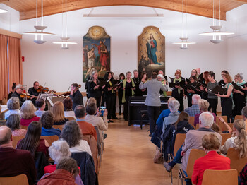 In einem Pfarrsaal sitzen viele Personen in Sitzreihen und hören einem Streichorchester am Ende des Raumes zu.