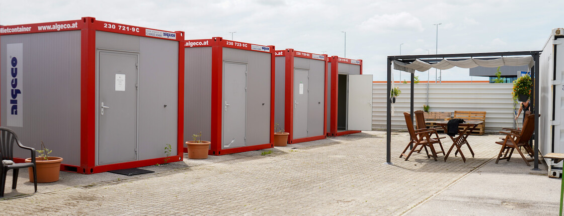 Caritas ZufluchtsDorf Mehrere rot-graue Container im Freien, gegenüber ein überdachter Sitzbereich.