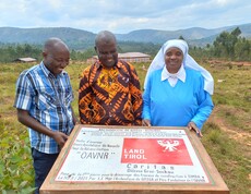 Drei lachende Personen – zwei Männer und eine Ordensschwester in blauem Habit – halten gemeinsam eine Projekttafel mit den Logos von Land Tirol und Caritas Diözese Graz-Seckau; im Hintergrund das fertiggestellte Gebäude des Ausbildungszentrums in Simba, Burundi.