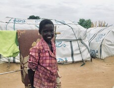 Lächelndes Kind in kariertem Hemd vor provisorischen Zelten im Camp Gumbo, Südsudan.