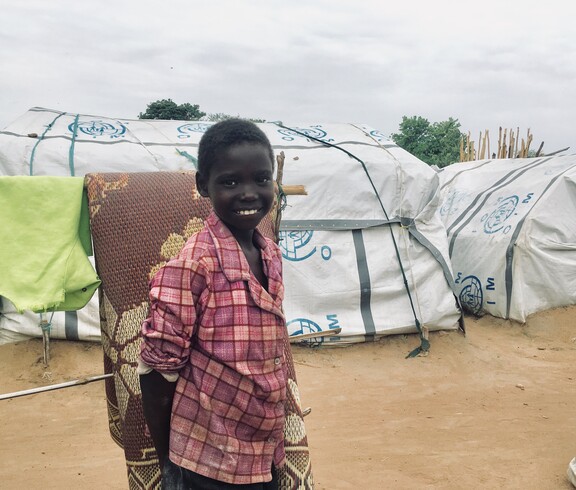 Lächelndes Kind in kariertem Hemd vor provisorischen Zelten im Camp Gumbo, Südsudan.