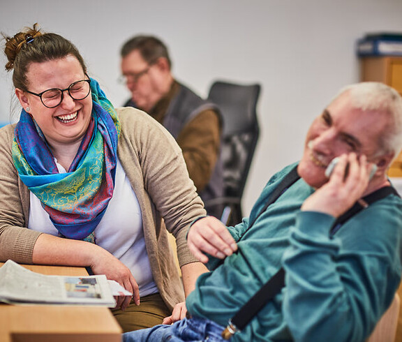 Frau und Mann lachen gemeinsam an einem Tisch, der Mann telefoniert in einer betreuten Umgebung