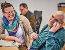 Frau und Mann lachen gemeinsam an einem Tisch, der Mann telefoniert in einer betreuten Umgebung