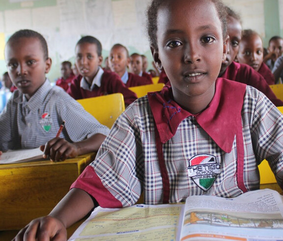 Ein Mädchen in der Schuluniform der Tiigo School blickt aufmerksam in die Kamera; im Hintergrund sitzen zahlreiche weitere Kinder in Uniform an gelben Schulbänken im voll besetzten Klassenraum.