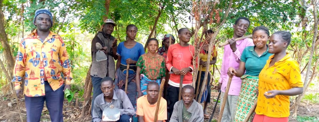 Eine Gruppe von Frauen und Männern steht gemeinsam mit Werkzeugen in einer bewaldeten ländlichen Umgebung.