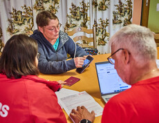 Eine Frau zeigt auf ihrem Smartphone Informationen, während sie mit anderen am Tisch sitzt. Die Gruppe bespricht Hilfeleistungen für Hochwasseropfer in Böheimkirchen. Ein Laptop und ein Reisepass sind auf dem Tisch sichtbar, was auf eine formale Beratung hinweist.