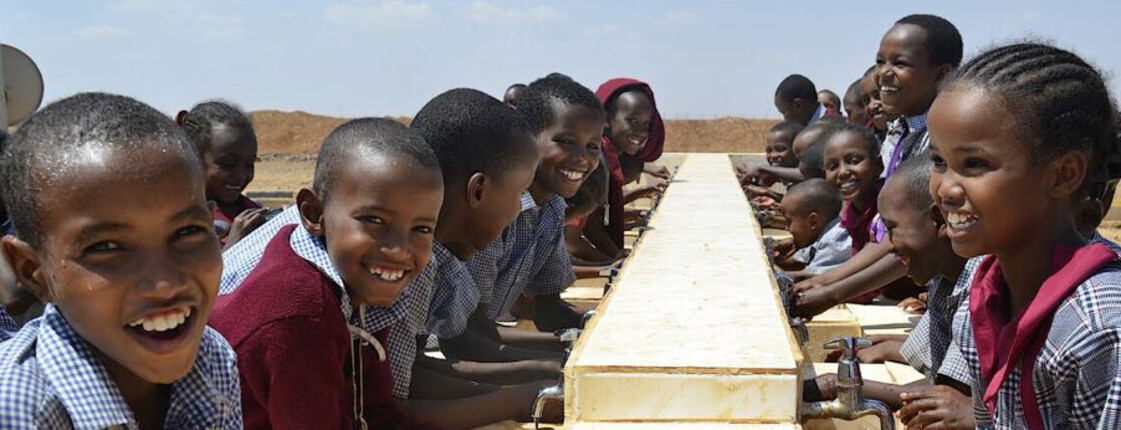 Eine Gruppe lachender Kinder in Schuluniform steht an einer langen Außen-Handwaschanlage aus hellem Stein in der trockenen Landschaft von Marsabit, Kenia; © PACIDA