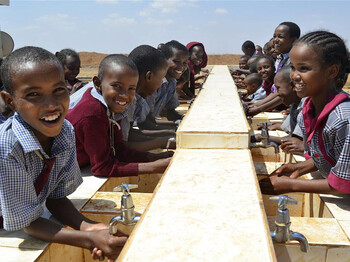 Eine Gruppe lachender Kinder in Schuluniform steht an einer langen Außen-Handwaschanlage aus hellem Stein in der trockenen Landschaft von Marsabit, Kenia; © PACIDA