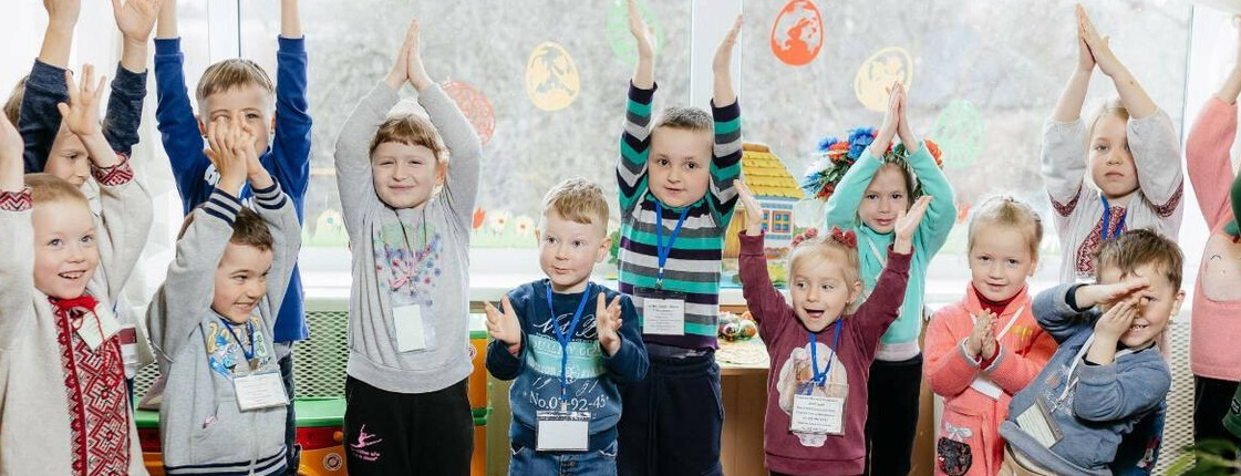 Eine Gruppe von kleinen Kindern steht in einem Halbkreis, sie lachen und werfen die Hände in die Höhe.