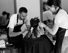 Zwei Personen führen gemeinsam eine Friseurbehandlung an einer sitzenden Person durch.