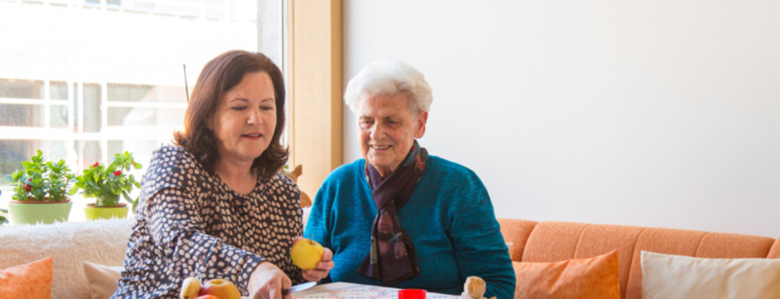 Eine Frau mittleren Alters sitzt mit einer alten Dame am Tisch. Die jüngere Dame hält einen Apfel in der Hand, den sie aufschneiden will.