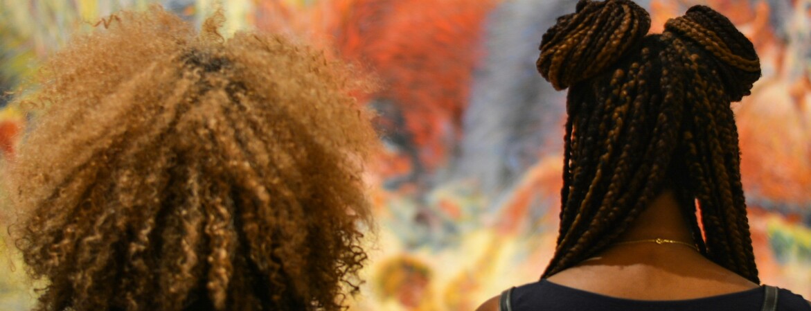 Zwei Frauen mit kreativen Frisuren, die Tanktops tragen, stehen in der Galerie vor einem bunten Gemälde.