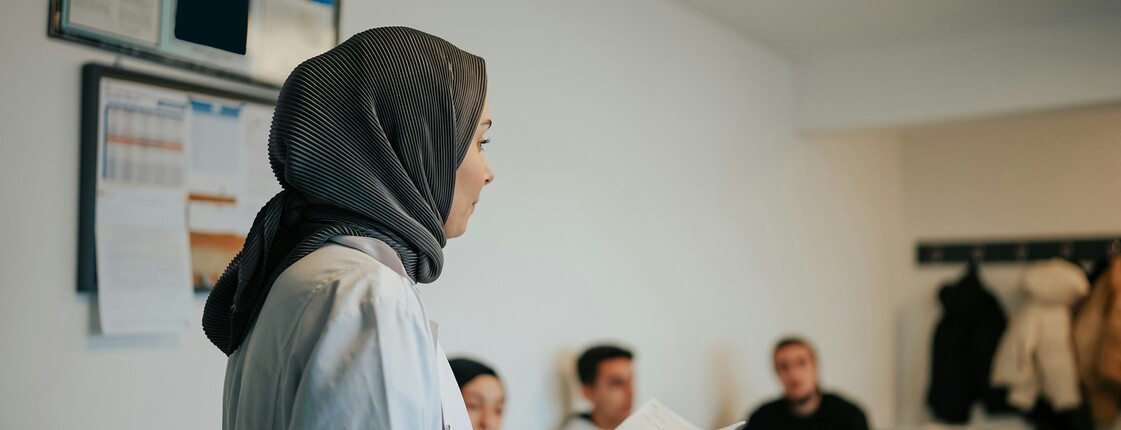 In einem Sprachkurs steht eine Frau mit Hijab vor den anderen Teilnehmer*innen und trägt etwas vor.