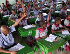Zahlreiche Kinder in der Schuluniform der Tiigo School sitzen an grünen Schulbänken und strecken enthusiastisch die Hände; auf den Tischen liegen Hefte und Bücher – ein lebhafter Unterricht im voll besetzten Klassenraum.