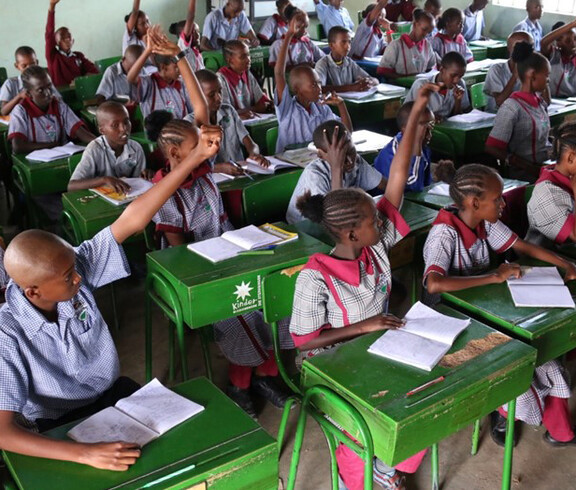 Zahlreiche Kinder in der Schuluniform der Tiigo School sitzen an grünen Schulbänken und strecken enthusiastisch die Hände; auf den Tischen liegen Hefte und Bücher – ein lebhafter Unterricht im voll besetzten Klassenraum.