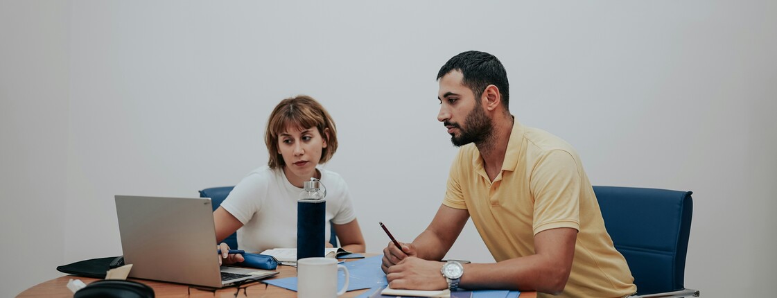 Eine Frau und ein Mann sitzen an einem runden Holztisch, auf dem Schreibunterlagen liegen, und schauen in einen Laptop.