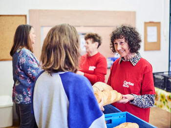 Eine freiwillige Mitarbeiterin der Caritas spricht lächelnd mit zwei Personen und verteilt Lebensmittel in einer Einrichtung.