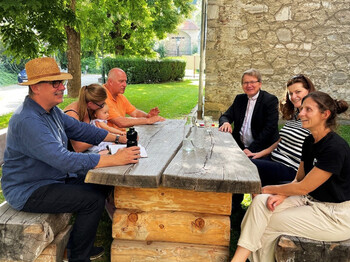 Menschen sitzen an einem Holztisch im schattigen Garten neben der Pfarrkirche und kommen zur Eröffnung der Schattenoase zusammen.