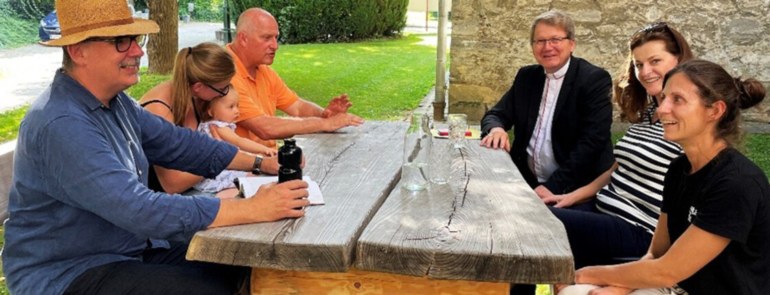 Menschen sitzen an einem Holztisch im schattigen Garten neben der Pfarrkirche und kommen zur Eröffnung der Schattenoase zusammen.