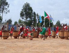Eine Gruppe von Männern in traditionellen rot-weißen Gewändern spielt synchron auf großen Holztrommeln bei einer feierlichen Zeremonie im Freien; im Hintergrund wehen die burundische Nationalflagge und weitere Fahnen.
