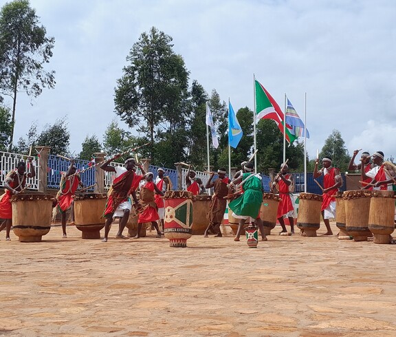 Eine Gruppe von Männern in traditionellen rot-weißen Gewändern spielt synchron auf großen Holztrommeln bei einer feierlichen Zeremonie im Freien; im Hintergrund wehen die burundische Nationalflagge und weitere Fahnen.