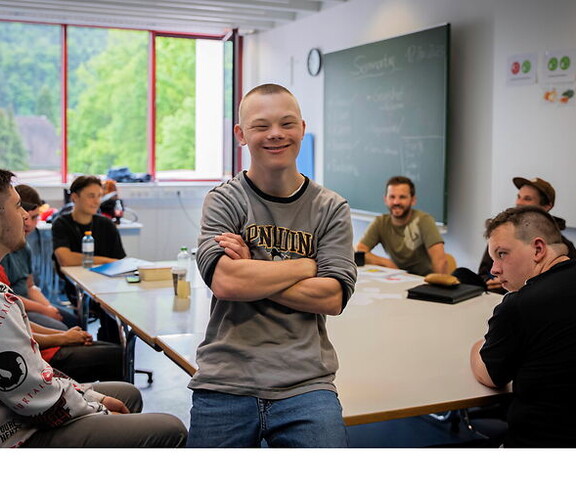 Junger Mann mit Behinderung steht lächelnd in einem Klassenraum mit anderen Jugendlichen