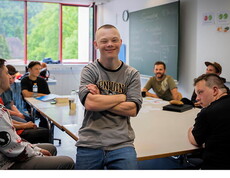 Junger Mann mit Behinderung steht lächelnd in einem Klassenraum mit anderen Jugendlichen