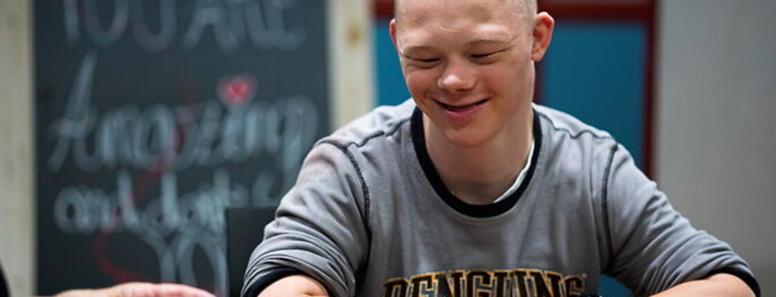 Junger Mann mit Behinderung sitzt lächelnd am Tisch in einer betreuten Alltagssituation