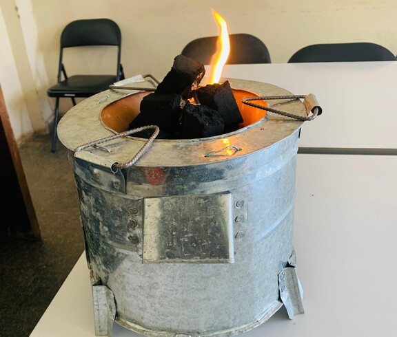 Im Projekt PERDURER kommen Energiesparöfen als saubere Kochalternative zum Einsatz  Metallofen mit brennenden Briketts auf einem Tisch.