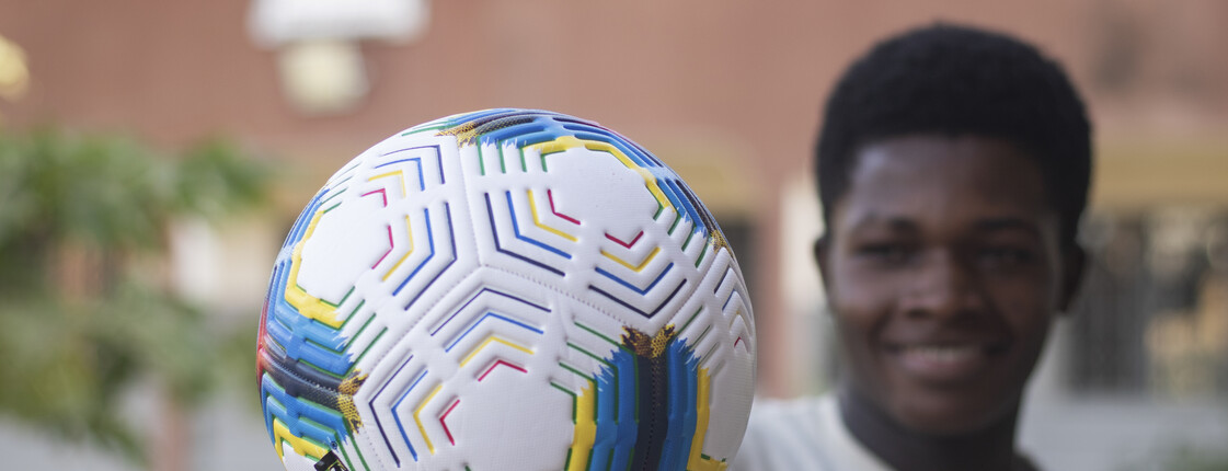 Nahaufnahme eines weißen Fußballs mit bunten Streifen, der von einem lächelnden Jugendlichen vor die Kamera gehalten wird.