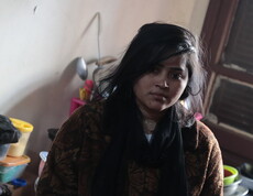 Frau sitzt in einem kleinen Raum neben Haushaltsgegenständen und Küchenutensilien.