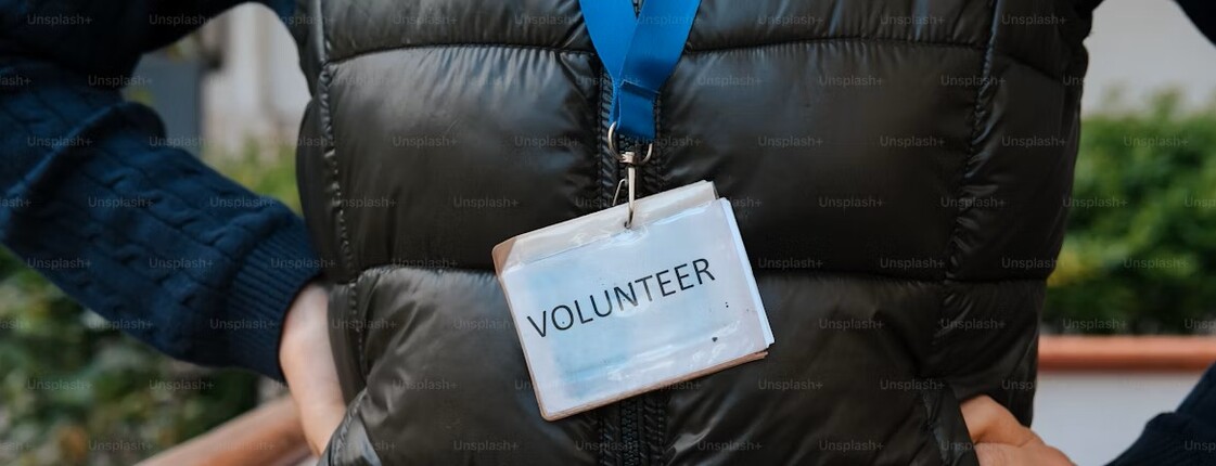 Eine Frau hat eine schwarze Steppweste an und trägt ein Schild mit der Aufschrift "Volunteer".