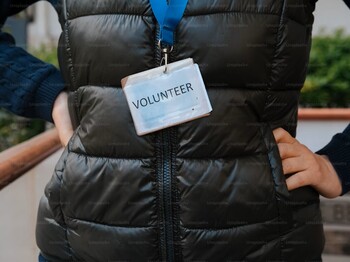 Eine Frau hat eine schwarze Steppweste an und trägt ein Schild mit der Aufschrift "Volunteer".