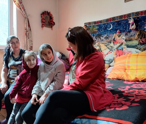 Zwei Mädchen und zwei Frauen sitzen auf einer Bettkante. Im Hintergrund ist ein bunter Wandteppich.
