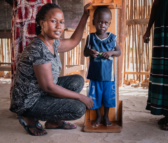 eine südsudanesische Frau misst die Größe eines Kleinkindes und blickt in die Kamera