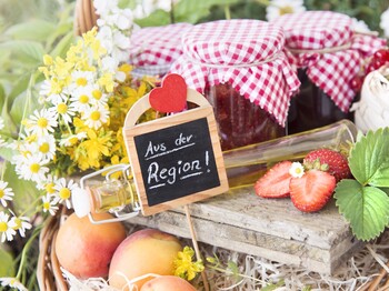 auf einem Holzbrett Marmeladengläser mit rot-weiß Karierten Tüchern auf den Deckeln daneben weiße und gelbe Blumen, Marillen, Erdbeeren aufgeschnitten + Blüte und Blatt dazu ein Holzsticker mit Herz, worauf steht aus der Region