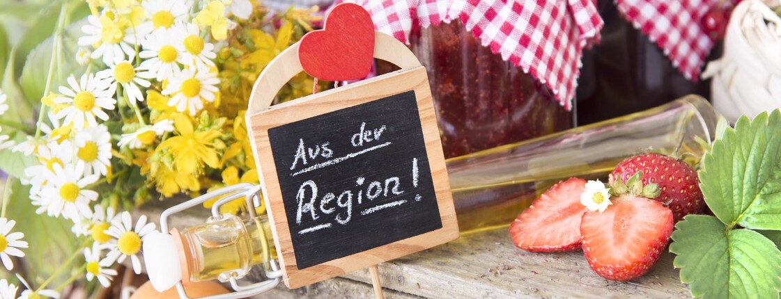 auf einem Holzbrett Marmeladengläser mit rot-weiß Karierten Tüchern auf den Deckeln daneben weiße und gelbe Blumen, Marillen, Erdbeeren aufgeschnitten + Blüte und Blatt dazu ein Holzsticker mit Herz, worauf steht aus der Region