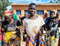 Eine burundische Frau trägt ein Ziegenkitz auf dem Arm und lächelt in die Kamera. Um sie stehen weitere Frauen. 