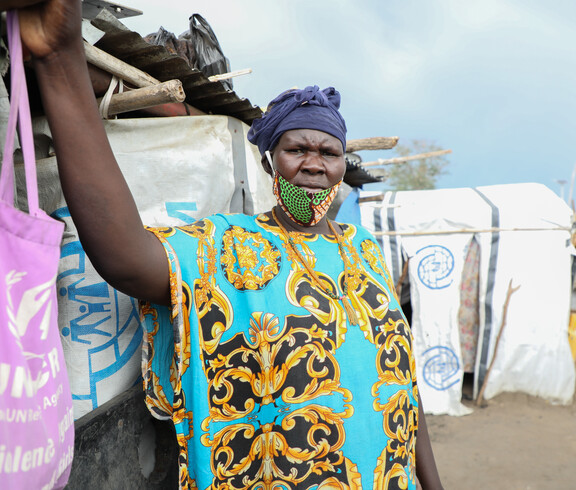 Porträt einer älteren Frau mit blauem Kopftuch und gemustertem Kleid vor provisorischen Unterkünften aus Planen und Holzstangen im Camp Gumbo, Südsudan