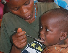 Nahaufnahme eines kleinen Kindes, das von einer erwachsenen Person mit einem Löffel gefüttert wird – Versorgung im Ernährungszentrum des Caritas-Projekts in Gitega, Burundi.