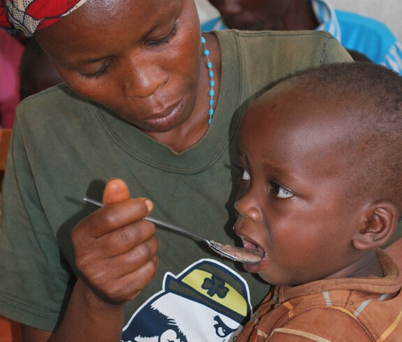 Nahaufnahme eines kleinen Kindes, das von einer erwachsenen Person mit einem Löffel gefüttert wird – Versorgung im Ernährungszentrum des Caritas-Projekts in Gitega, Burundi.