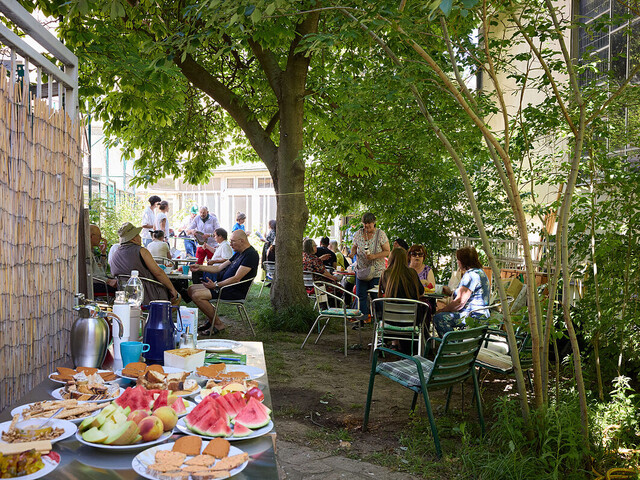 Mehrere Menschen sitzen im Schatten der Bäume zusammen im Freien, im Vordergrund ein Buffet mit frischen Früchten und Getränken.