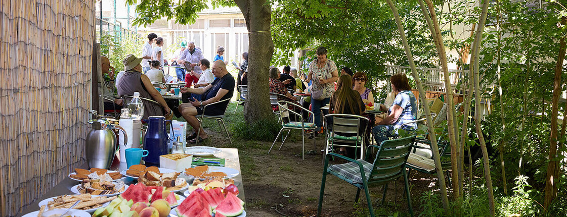 Mehrere Menschen sitzen im Schatten der Bäume zusammen im Freien, im Vordergrund ein Buffet mit frischen Früchten und Getränken.