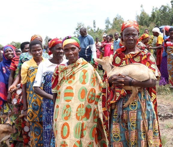 Burundische Frauen stehen aufgereiht in feierlichen Kleidern, die Frau am linken Rand trägt eine Ziege in den Armen. 