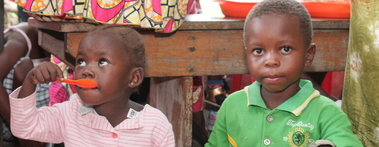 Ein kleines Mädchen und ein Junge sitzen auf dem Boden und essen Brei mit Löffeln in einer Ernährungsstation